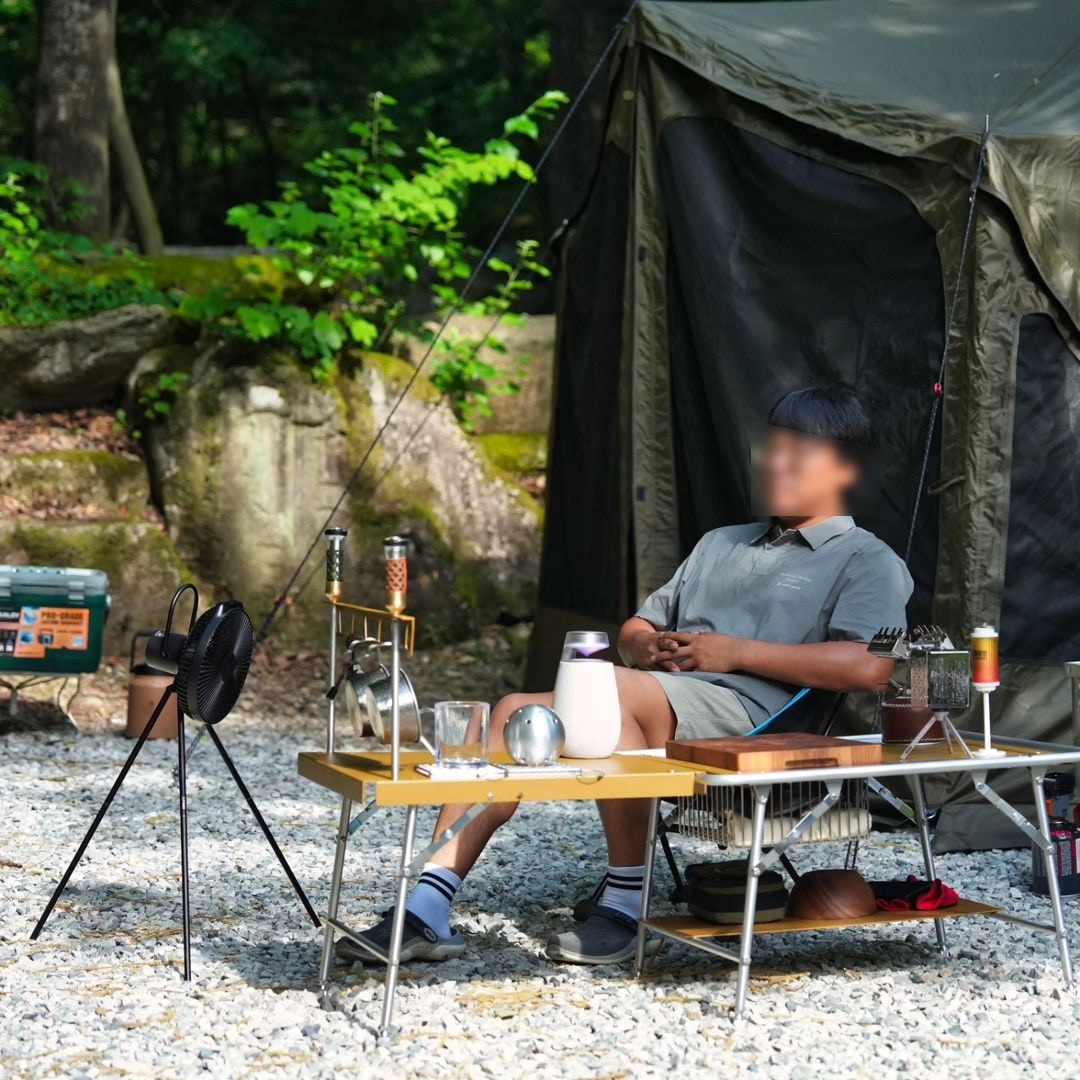 캠핑 테이블 위에 엑스붐 360와 여러 캠핑용 기구들이 배치되어 있고, 한 남성이 캠핑의자에 앉아 있음. 배경에는 녹음이 우거진 숲과 텐트가 보임.
