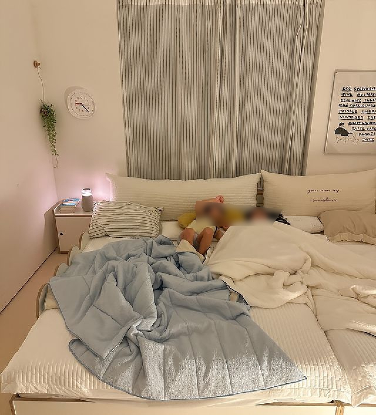 커튼이 드리워진 침실, 작은 테이블 위에 점등된 엑스붐 360가 배치됨. 침대에 흰색 및 파란색 침구가 깔려 있으며, 벽에는 시계와 그림이 걸려 있음. 침대 위에는 사람들이 자고 있음.