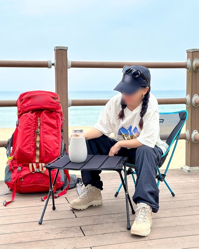 푸른 바다와 하늘가 뒤로 보이는 바닷가 데크에서 캠핑 의자에 앉아 테이블 위에 있는 엑스붐 360을 만지고 있는 여성. 옆에는 빨간 베낭이놓여있음.