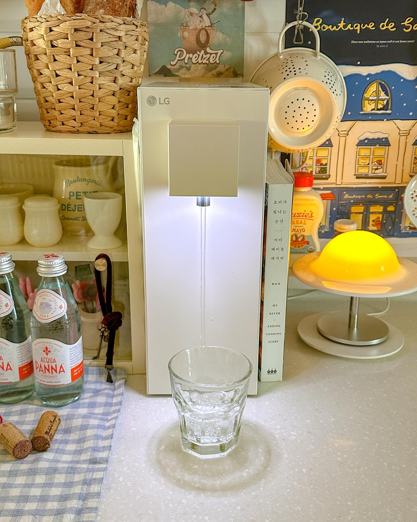 LG 정수기가 조리대 위에서 물을 컵에 따르고 있으며, 주변에는 병음료, 주방 장식품, 조명이 아늑한 분위기를 연출하고 있음