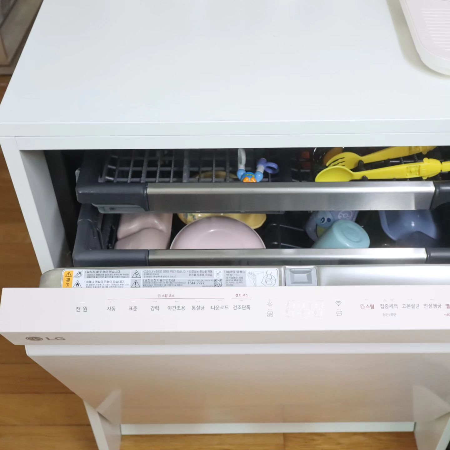 흰색 LG 식기세척기의 내부, 그릇과 장난감, 숟가락 등이 배치된 모습, 식기세척기 하단에는 설정 버튼과 디스플레이가 보임