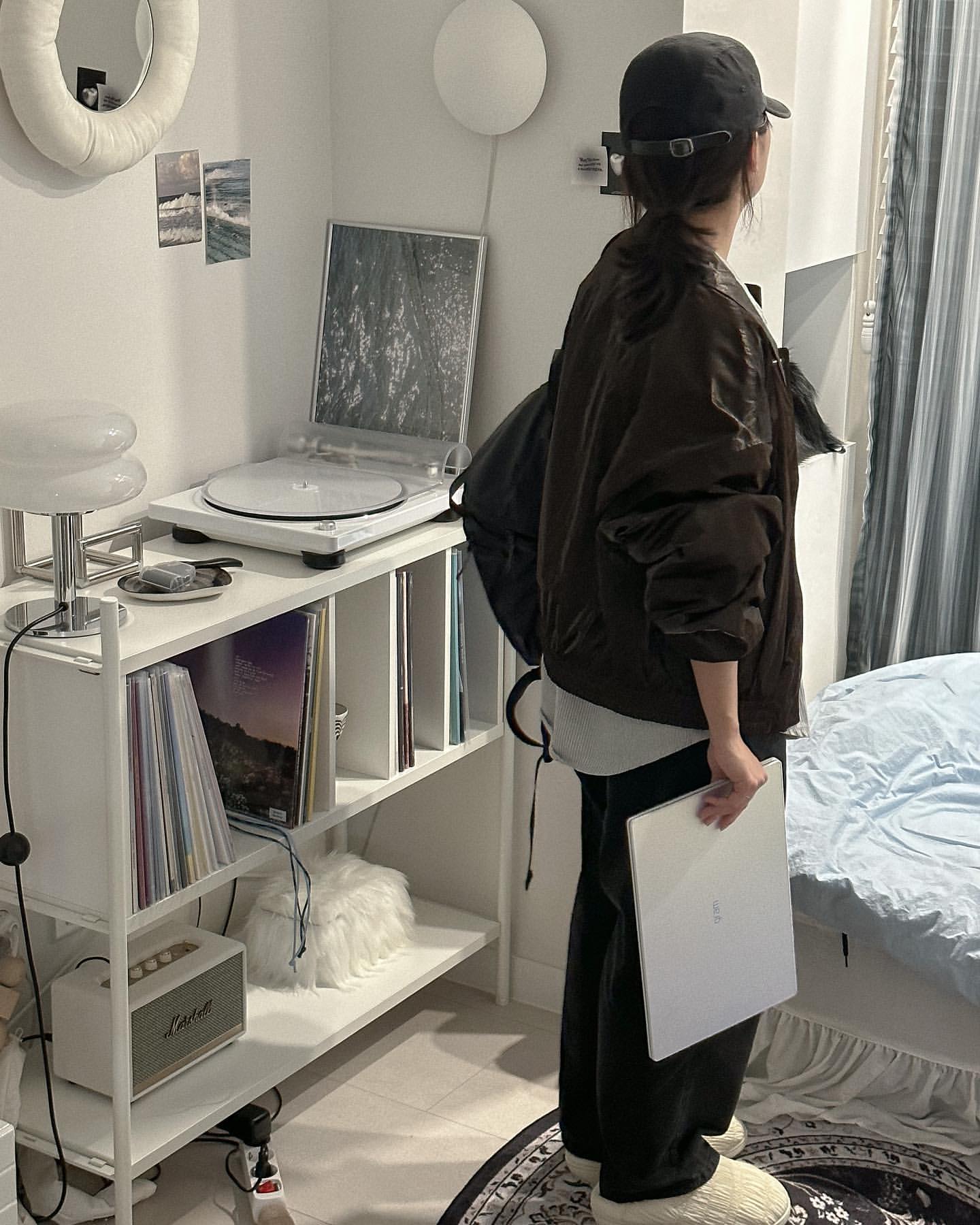 검은색 모자와 점퍼를 입은 여성이 LG 그램 Style 노트북을 들고 책장 앞에 서 있으며 책장 위에는 턴테이블과 다양한 앨범이 놓여 있는 모습.