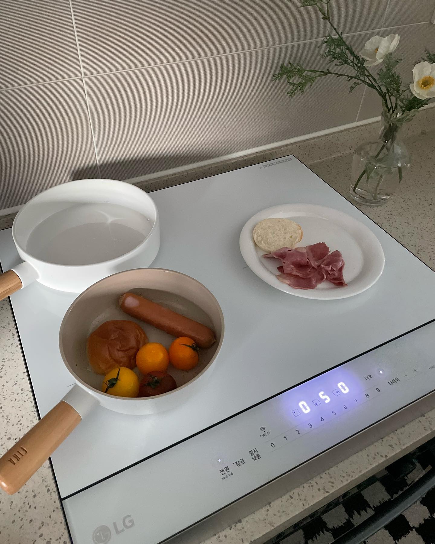 조리 중인 인덕션 위 냄비에 소시지와 토마토 등이 담겨 있고, 옆에 햄과 빵이 담긴 접시가 놓여 있는 모습.