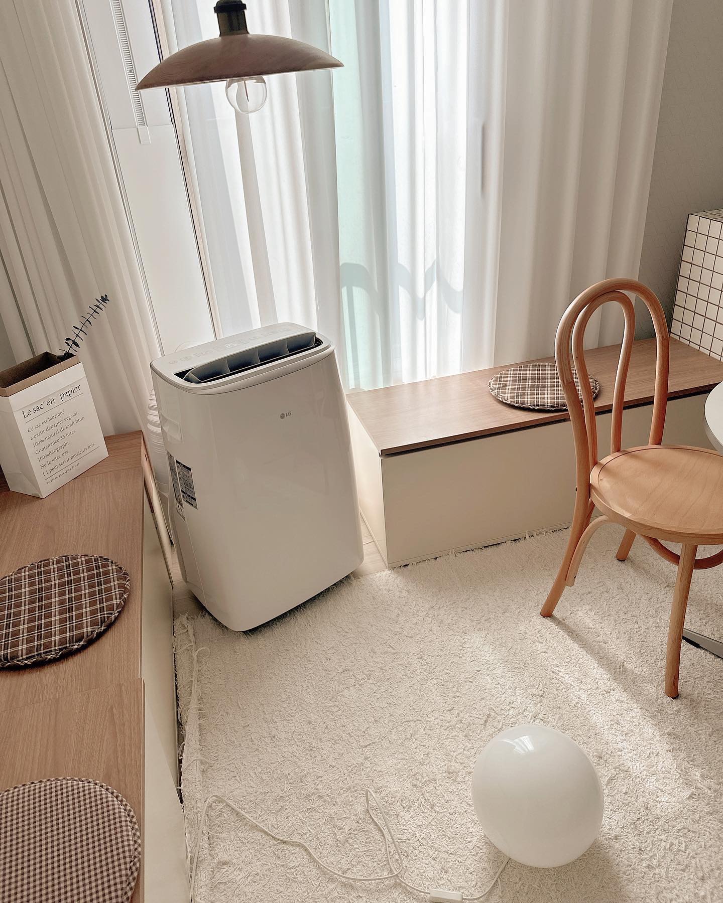LG햇살이 비추는 공간에 이동식 에어컨과 나무 의자, 체크 패턴의 방석이 놓인 벤치, 흰색 러그와 둥근 조명이 배치된 따뜻한 분위기의 코너
