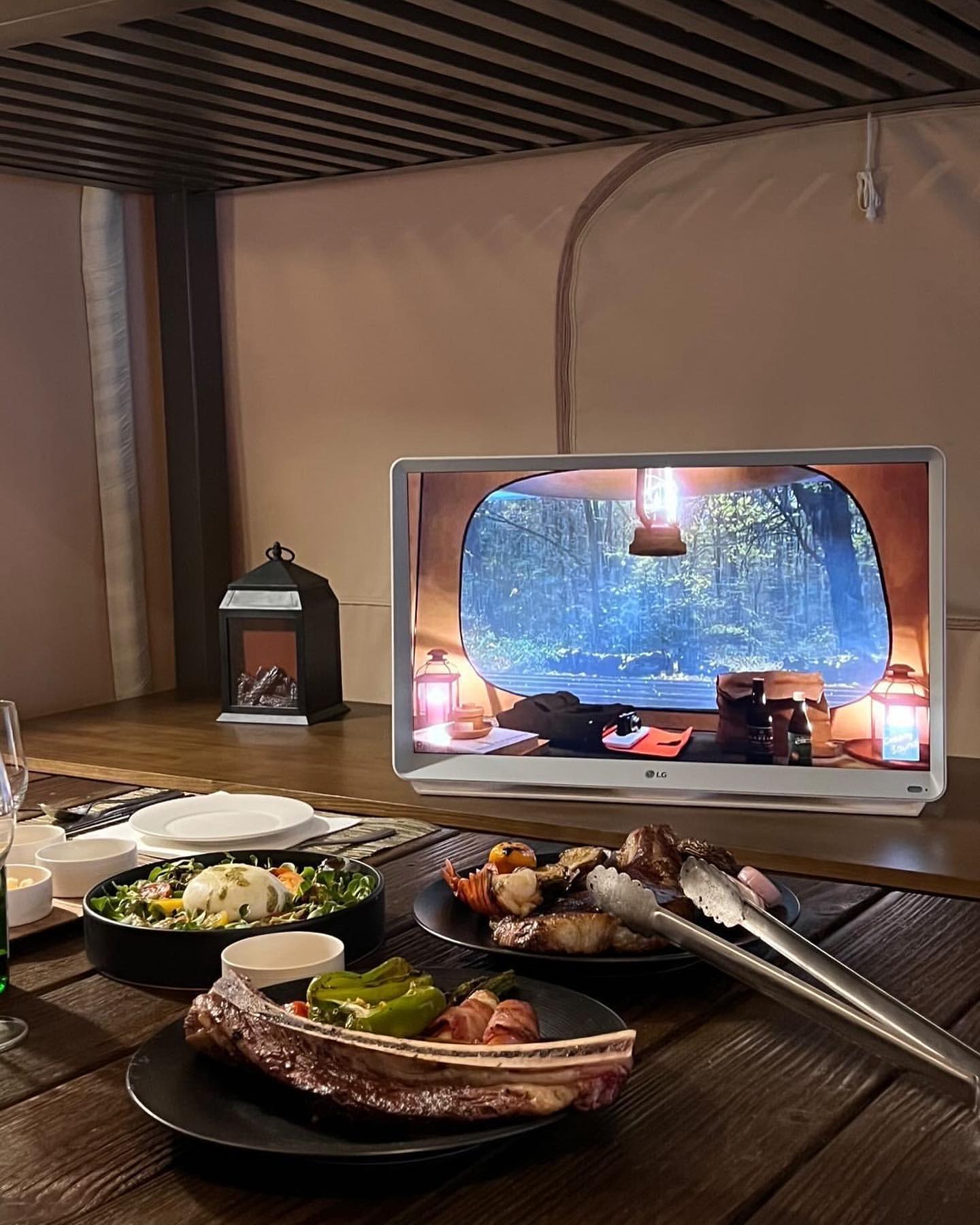 캠핑 텐트 내부에 LG 룸앤 TV가 설치되어 있으며, 앞에는 음식들이 놓여 있는 모습.