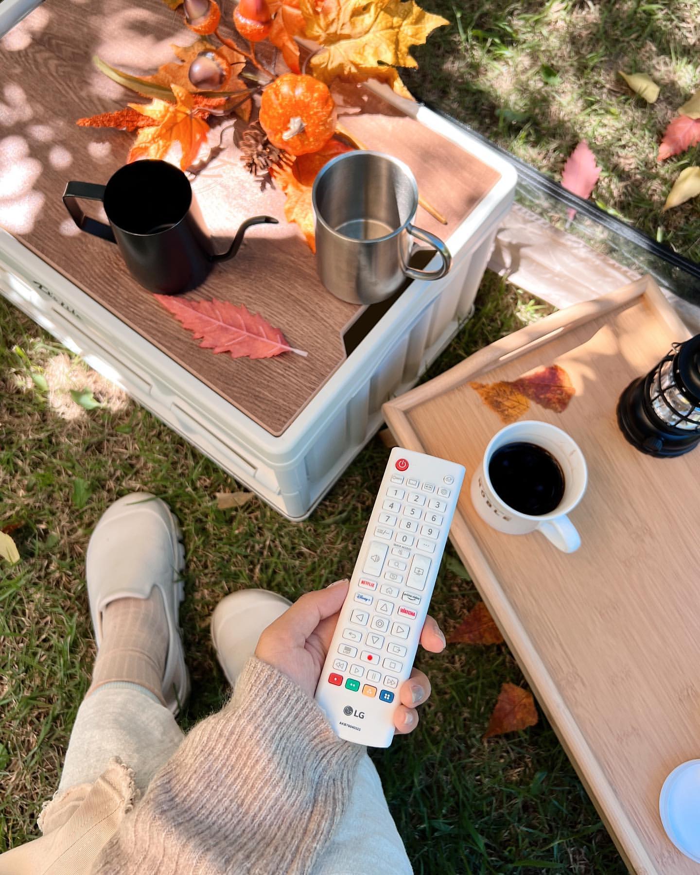 LG 룸앤 TV 리모컨을 들고 있는 손 이미지와 근처에는 나무 받침 위에 컵들이 놓여 있는 모습.