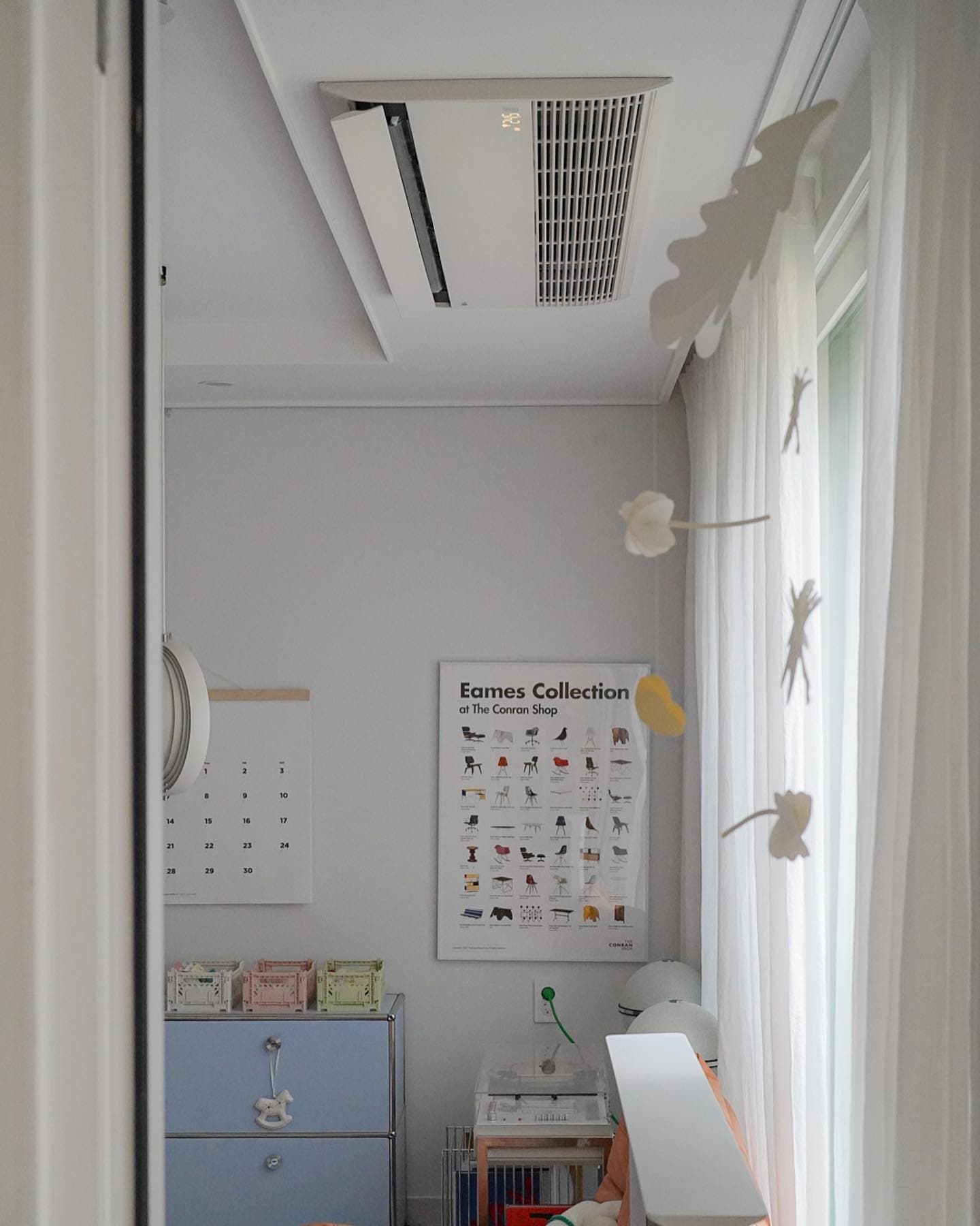 천장에 설치된 시스템 에어컨과 창가의 밝은 커튼, 깔끔하게 정리된 서랍장과 포스터로 꾸며진 정돈된 실내 공간