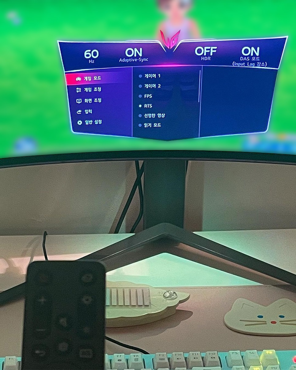 LG 울트라와이드 게이밍 모니터 화면에 게임 모드 및 설정 메뉴가 표시되고, 책상 위에 리모컨이 놓인 모습.