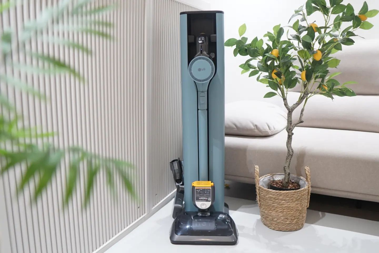 거실 공간에 놓인 무선 청소기 충전 스테이션. 청소기는 세련된 파란색 디자인으로, 옆에는 작은 화분에 심어진 인조 나무와 베이지색 소파가 보임