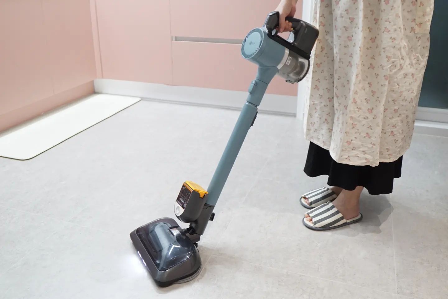 사람이 무선 청소기를 사용하여 부엌 바닥을 청소하는 모습. 청소기는 파란색 본체에 투명한 먼지통이 있으며, 바닥 청소 헤드에는 LED 조명이 켜져 있다. 사용자는 꽃무늬 원피스와 줄무늬 슬리퍼를 착용하고 있음