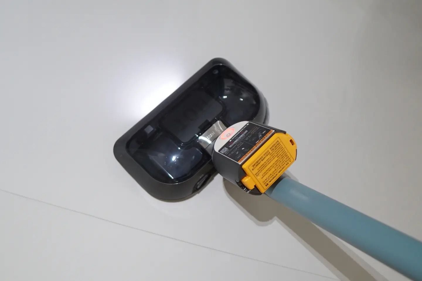 밝은 바닥 위에서 작동 중인 무선 청소기. 청소기 헤드 부분의 LED 조명이 켜져 있으며, 상단에는 스팀 기능을 활성화하는 버튼과 경고 라벨이 부착되어 있음