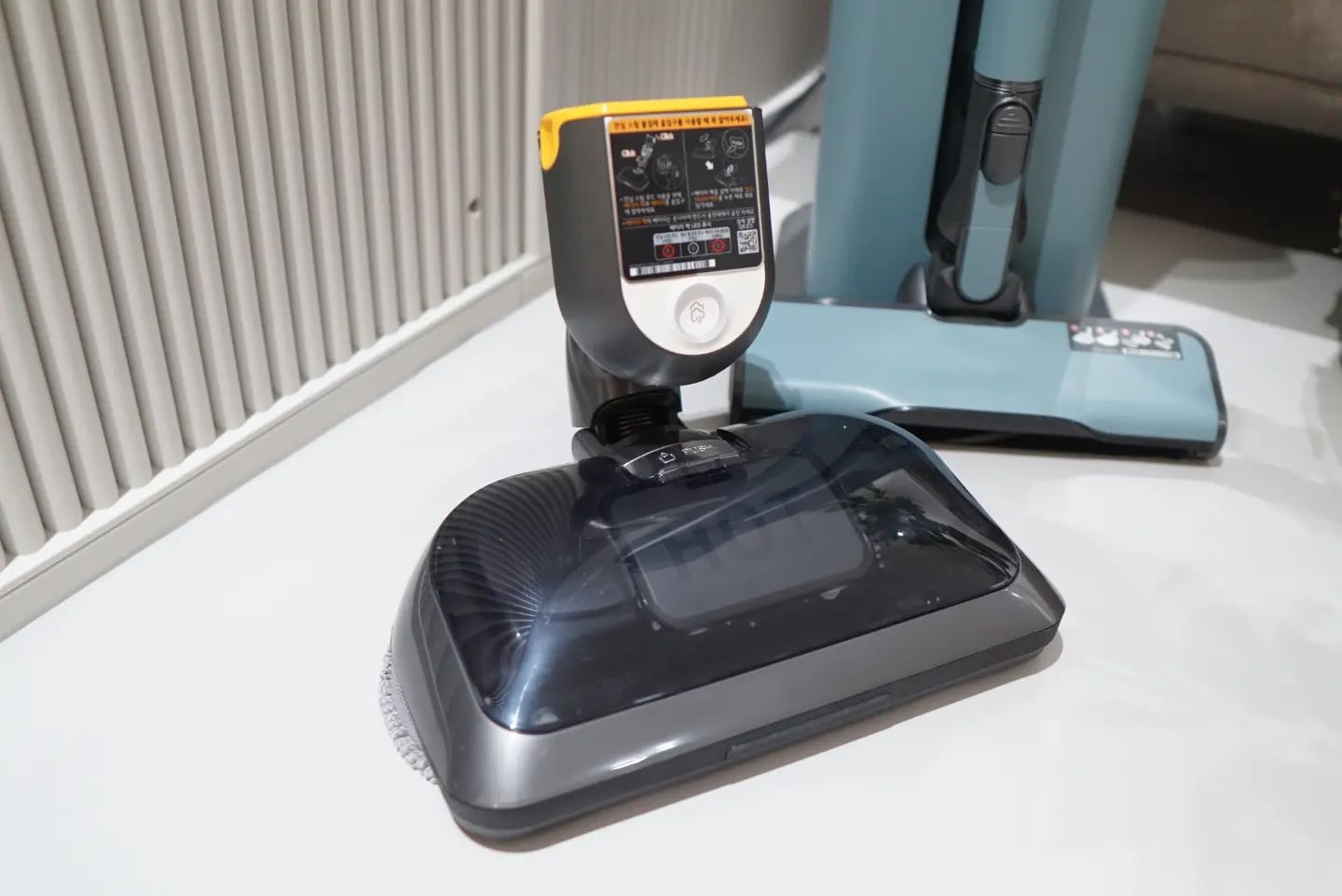 분리된 무선 청소기 헤드가 바닥에 놓여 있으며, 상단에는 스팀 기능을 나타내는 버튼과 주의사항이 적힌 경고 라벨이 부착되어 있음