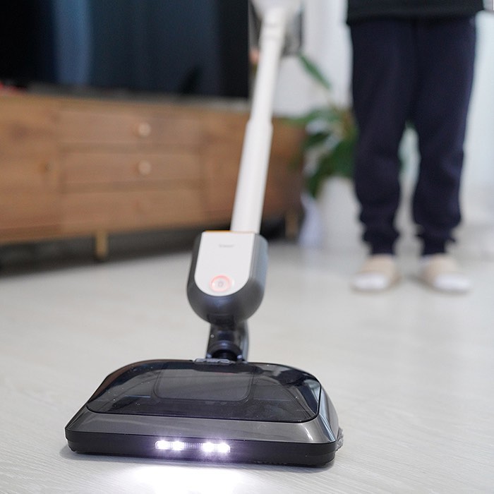 LED 조명이 켜진 무선 물걸레 청소기가 밝은 바닥을 청소 중인 모습. 청소기를 조작하는 사람의 다리와 슬리퍼가 보이며, 배경에는 나무 수납장이 놓여 있음