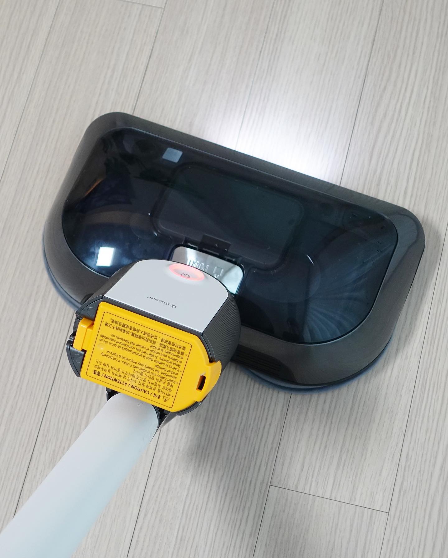 나무 바닥을 청소 중인 무선 물걸레 청소기, 투명한 청소 헤드와 스팀 기능 버튼이 있으며, 노란색 주의사항 라벨이 부착되어 있음