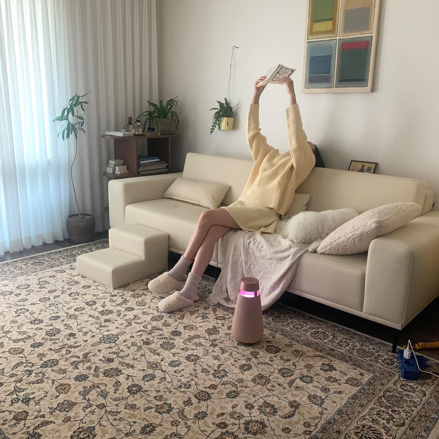 베이지색 소파에 앉아 책을 들고 있는 사람과 소파 옆에 누워 있는 강아지, 그리고 화려한 패턴의 러그와 엑스붐, 식물이 배치된 아늑한 거실