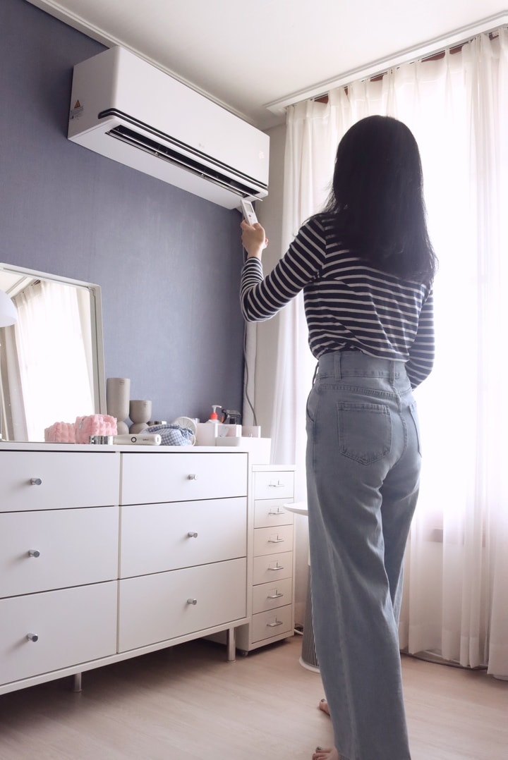여성이 무선 리모컨으로 벽걸이 에어컨을 조작하고 있음. 배경에는 조명, 시계, 화장품과 거울이 정리된 흰색 서랍장과, 흰색 철제 서랍장이 배치되어 있고, 가려진 커튼 사이로 자연광이 들어옴.