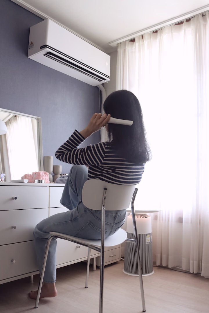 남색 톤 벽의 방에서 여성이 의자에 앉아 에어컨 리모컨을 들고 있음. 배경에는 화장품과 거울이 정리된 흰색 서랍장과 흰색 철제 서랍장이 배치됨. 테이블형 공기청정기가 있으며, 가려진 커튼 사이로 자연광이 들어옴.