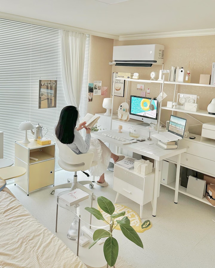 흰색 블라인드가 내려지고 커튼이 묶인 화사한 방 안, 작동 중인 벽걸이 에어컨 아래에서 여성이 의자에 앉아 책을 읽음. 책상 위엔 디지털 기기와 아이템들이 정리되어 있으며, 침대, 전등, 노란 수납장, 초록 식물이 조화를 이룸.