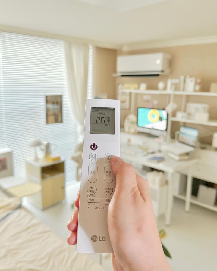 에어컨 리모컨을 들고 26°C로 설정된 온도를 조정하는 손, 배경에는 침구와 작업 공간의 모니터, 책상, 식물들이 배치돼 있고, 창가에 화이트 커튼과 블라인드가 있음. 