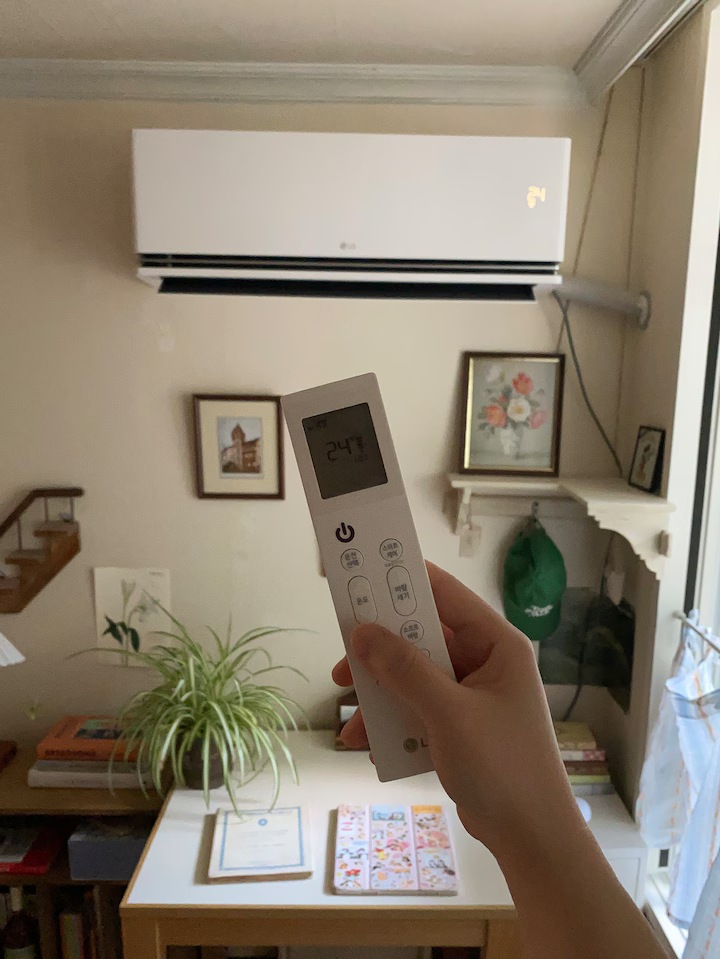 벽걸이 에어컨 화면에 24°C가 표시됨. 손에 리모컨을 들고 있음. 배경에는 책상 위의 식물, 책, 문구류가 보이며, 벽에는 액자들, 벽 선반에는 초록색 모자가 걸려있음.