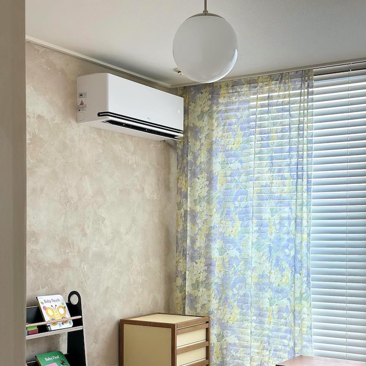 벽걸이 에어컨이 있는 아기방, 창문에는 블라인드가 내려져 있고, 꽃무늬 커튼이 걸려 있음. 책이 놓인 선반과 서랍장이 있음.