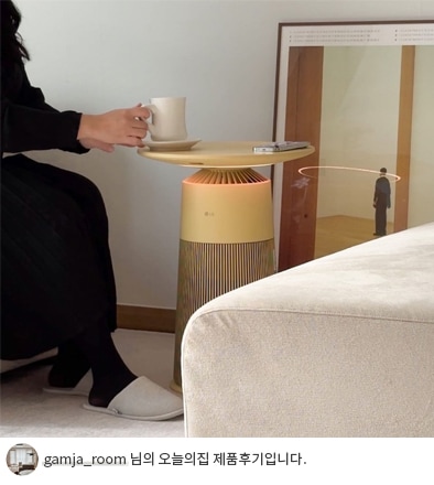 검정색 옷을 입은 여성이 LG 퓨리케어 오브제컬렉션 에어로퍼니처 상단을 테이블로 사용해 컵을 올려놓고 앉아 있는 모습과 하단에 'gamja_room 님의 오늘의집 제품후기입니다.' 텍스트가 포함된 모습.