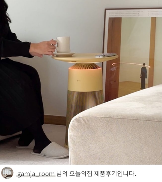 검정색 옷을 입은 여성이 LG 퓨리케어 오브제컬렉션 에어로퍼니처 상단을 테이블로 사용해 컵을 올려놓고 앉아 있는 모습과 하단에 'gamja_room 님의 오늘의집 제품후기입니다.' 텍스트가 포함된 모습.