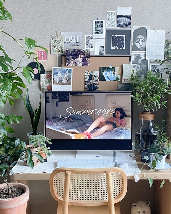 LG 일체형 PC가 책상 위에 놓여 있으며, PC 화면에는 'Summer 1983'라는 텍스트와 함께 여성 모델의 이미지가 표시됨. 책상 주변에는 다양한 크기의 화분과 식물들이 배치되어 있고, 벽에는 사진과 포스터들이 빼곡히 붙어 있어 갤러리 같은 분위기를 연출.