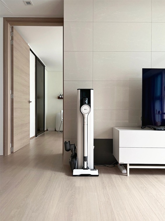 LG 코드제로 오브제컬렉션 A9S 무선청소기가 거실 벽 한쪽에 세워져 있는 모습. 세련된 화이트와 블랙 컬러의 본체가 깔끔하게 정리되어 있으며, 옆에는 TV와 콘솔이 배치되어 현대적이고 실용적인 인테리어를 보여줌.