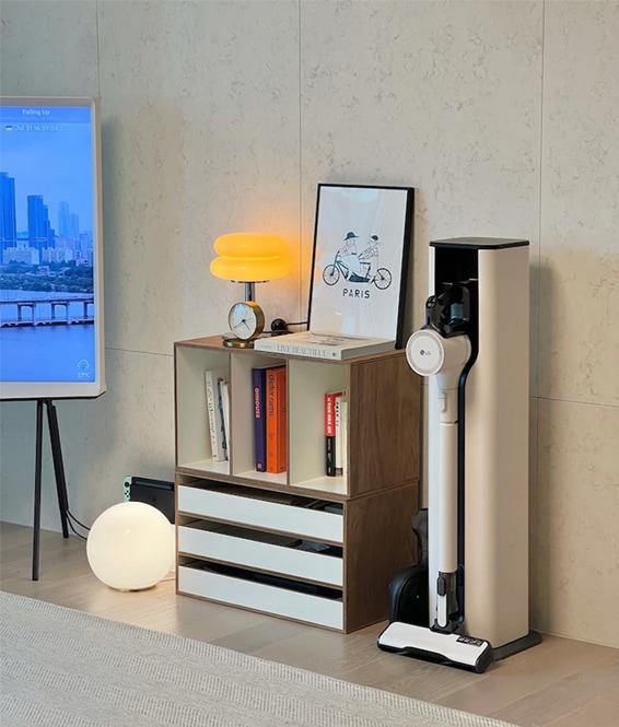 거실 선반 옆에 LG 코드제로 오브제컬렉션 A9S 무선청소기가 충전 스탠드에 깔끔하게 배치되어 있는 모습. 심플한 인테리어와 조화를 이루고 있습니다.