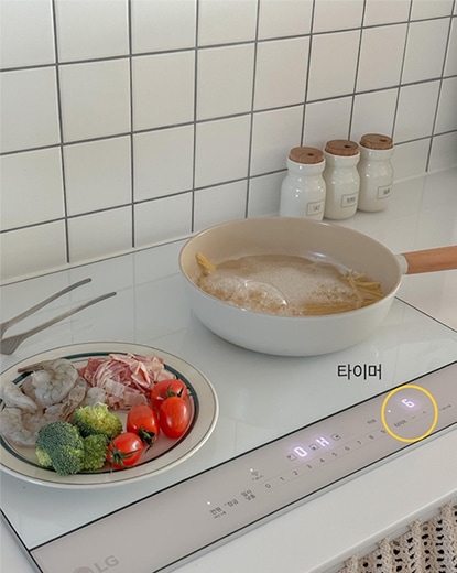 LG 디오스 오브제컬렉션 인덕션 위에서 요리가 진행 중인 모습. 냄비에는 국물이 끓고 있으며, 옆에는 야채와 새우가 담긴 팬이 놓여 있음. 인덕션의 터치 디스플레이가 작동 중이며, 주방의 화이트톤 인테리어와 조화를 이루고 있음.
