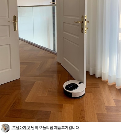 G 코드제로 오브제컬렉션 M9 로봇청소기가 방 문턱을 넘어 이동하는 모습. 배경에는 화이트 도어와 커튼으로 구성된 밝은 인테리어가 보임. 하단 텍스트: '호텔마가렛 님의 오늘의집 제품후기입니다.'