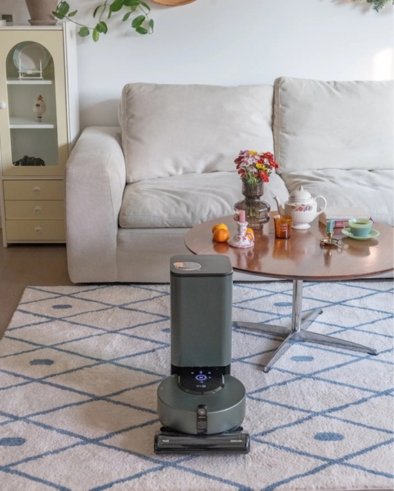 LG 코드제로 오브제컬렉션 R9 로봇청소기가 거실에 충전 거치대와 함께 배치된 모습. 거실에는 소파와 테이블 위에 꽃병과 음료가 놓여 있으며, 로봇청소기의 세련된 디자인이 깔끔한 인테리어와 조화를 이루고 있습니다.