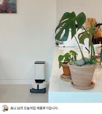 LG 코드제로 오브제컬렉션 R9 로봇청소기가 거실에서 작동 중인 모습. 주변에는 초록 식물 화분과 밝은 인테리어가 어우러져 깔끔하고 자연스러운 공간을 연출. 하단에는 '츄니 님의 오늘의집 제품후기입니다.'라는 텍스트가 포함되어 있음.