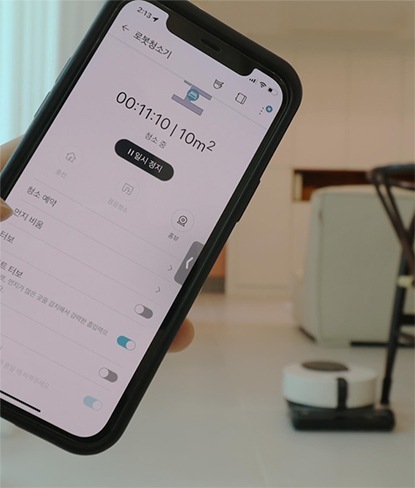 LG 코드제로 오브제컬렉션 R9 로봇청소기의 전용 앱 화면이 스마트폰에 표시된 모습. 청소 상태, 청소 시간 등의 정보가 표시되어 있으며, 뒤에는 로봇청소기가 거실에서 대기 중인 모습이 보임.