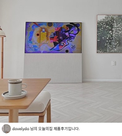 거실 테이블 위에 놓인 커피잔과 함께, 벽에 설치된 LG 올레드 오브제컬렉션 Easel TV에서 추상적인 예술 작품이 화면에 표시된 모습. 오른쪽 벽에는 꽃 그림 액자가 걸려 있으며, 아래에는 'dovelydo 님의 오늘의집 제품후기입니다.'라는 텍스트가 포함되어 있음.