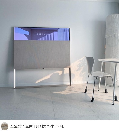 LG 올레드 오브제컬렉션 Easel TV가 하얀 벽을 배경으로 설치된 모습. 화면 상단에는 시간 '4:16 PM'이 표시되어 있음. 오른쪽 하단에는 의자와 작은 테이블이 보이며, 하단 텍스트는 '량쯔 님의 오늘의집 제품후기입니다.'라고 쓰여 있음
