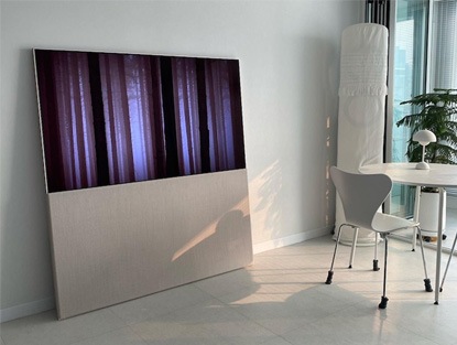 밝은 햇살이 비추는 거실에 배치된 LG 올레드 오브제컬렉션 Easel TV의 모습. TV 화면에는 은은한 색상의 이미지가 디스플레이되어 있고, TV 옆에는 자연광이 들어오는 커튼이 설치된 창문이 보임.