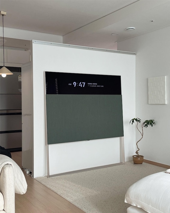 거실에 설치된 LG 올레드 오브제컬렉션 Easel TV. 흰색 벽면과 심플한 인테리어에 어우러져 있으며, 바닥에는 깔끔한 카펫이 깔려 있음. 옆에는 작은 화분이 놓여 있고, 전체적으로 차분한 분위기를 연출함.