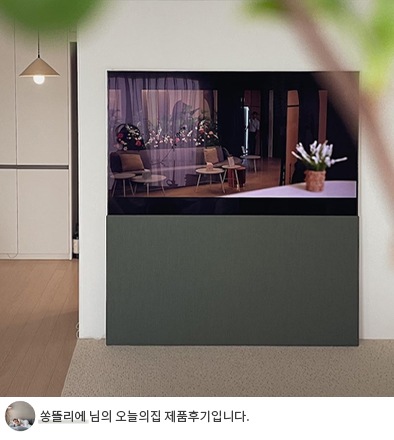 LG 올레드 오브제컬렉션 Easel TV 하단에 회색 패널이 세워져 있는 모습. TV 아래쪽에는 '쏭뜰리에 님의 오늘의집 제품후기입니다.'라는 텍스트가 포함되어 있음. TV는 실내 공간에서 예술 작품처럼 보이며, 모던한 디자인이 돋보임.