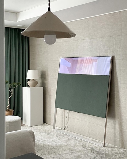 LG 올레드 오브제컬렉션 Easel TV가 다른 각도에서 촬영된 모습. 벽면과 가까운 위치에 배치되어 있으며, TV 뒷편으로는 녹색 커튼과 간접 조명이 있는 차분한 분위기의 실내 공간이 연출됨. TV는 예술적인 인테리어의 일부처럼 자연스럽게 어우러짐.