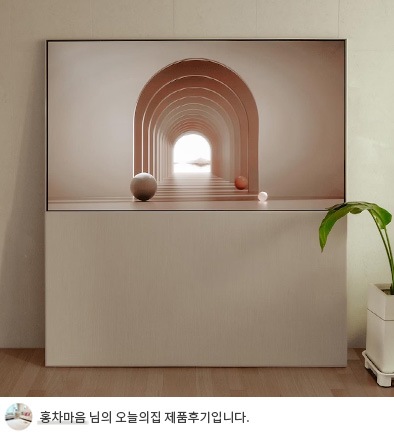 LG 올레드 오브제컬렉션 Easel TV의 무빙 커버가 내려간 상태로, 화면에 아치형 터널과 공이 보이는 감각적인 배경 이미지가 출력되어 있음. TV가 거실 벽에 설치되어 있고, 옆에는 녹색 화분이 배치되어 깔끔하고 현대적인 분위기를 연출. 하단에는 '홍차마음 님의 오늘의집 제품후기입니다.'라는 텍스트가 포함되어 있음.