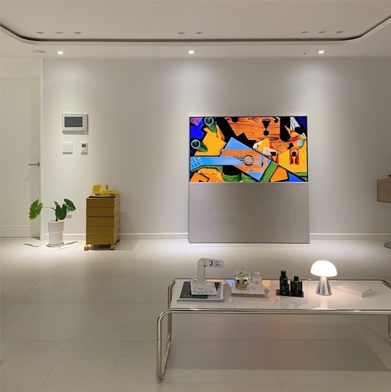 LG 올레드 오브제컬렉션 Easel TV가 거실 벽 중앙에 설치된 모습. TV 화면에는 화려한 색상의 추상화 이미지가 표시되어 있으며, 주변에는 화이트톤의 가구와 조명이 배치되어 깔끔하고 현대적인 거실 인테리어를 보여줌.