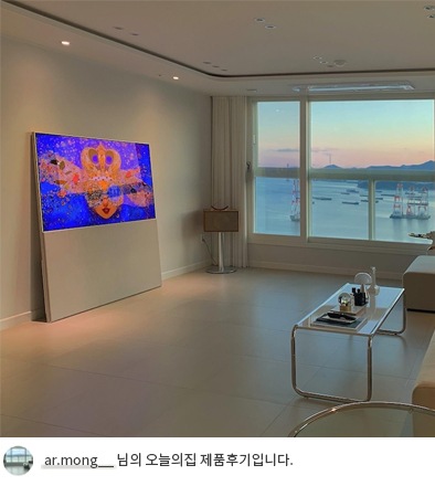 LG 올레드 오브제컬렉션 Easel TV가 넓은 거실 한쪽에 설치된 모습. TV 화면에는 파란색과 노란색이 조화를 이루는 이미지를 표시하고 있으며, 옆에는 창밖으로 보이는 석양과 바다 풍경이 고급스러운 분위기를 연출. 하단에는 'ar.mong__ 님의 오늘의집 제품후기입니다.'라는 텍스트가 포함되어 있음.
