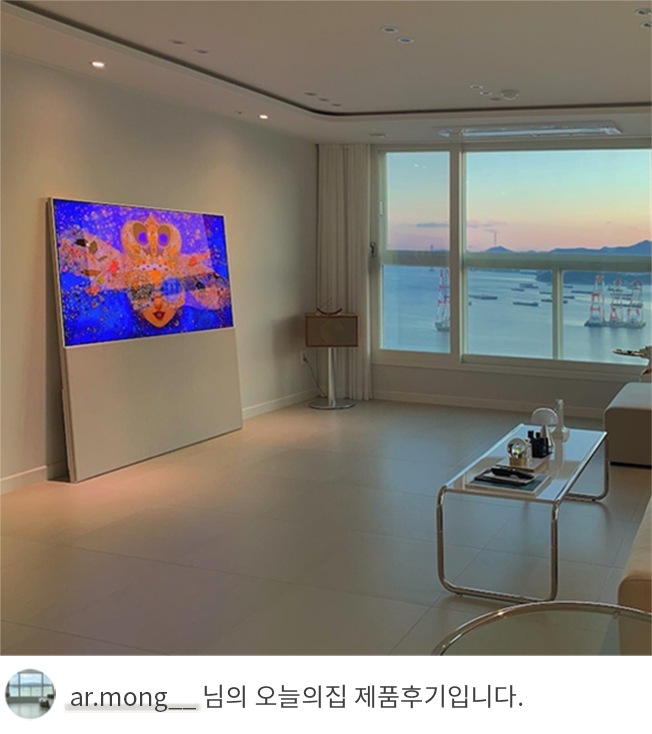 LG 올레드 오브제컬렉션 Easel TV가 넓은 거실 한쪽에 설치된 모습. TV 화면에는 파란색과 노란색이 조화를 이루는 이미지를 표시하고 있으며, 옆에는 창밖으로 보이는 석양과 바다 풍경이 고급스러운 분위기를 연출. 하단에는 'ar.mong__ 님의 오늘의집 제품후기입니다.'라는 텍스트가 포함되어 있음. 