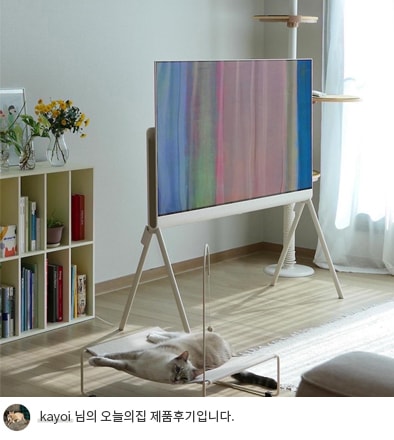 LG 올레드 오브제컬렉션 Posé TV가 나무 바닥 위에 세워져 있으며, 화면에는 추상적인 파스텔 톤의 이미지가 표시됨. 아래에는 고양이가 누워있는 모습이 보이며, 옆에는 책장이 배치되어 깔끔하고 모던한 거실 분위기를 연출. 하단에는 'kayoi 님의 오늘의집 제품후기입니다.'라는 텍스트가 포함되어 있음.