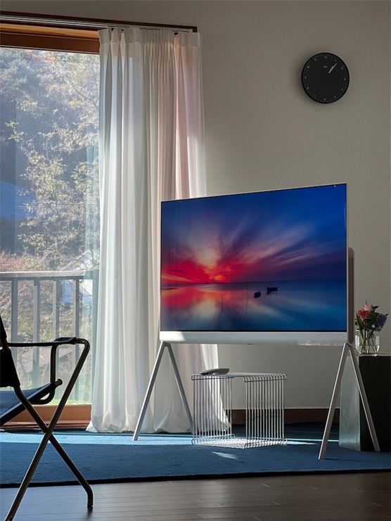 모던한 디자인의 거실에 LG 올레드 오브제컬렉션 포제(Posé) TV가 창가 옆에 배치되어 있으며, 벽시계와 의자가 함께 배치되어 세련된 인테리어를 완성하고 있습니다.