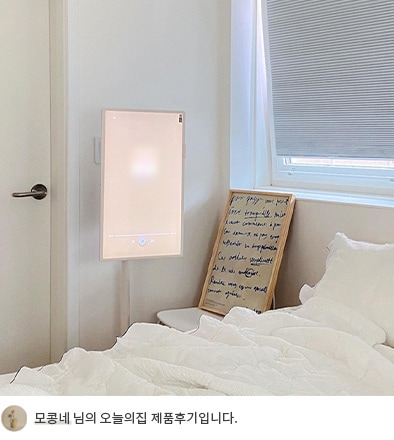 침실 옆에 설치된 LG 스탠바이미가 밝은 화면을 표시하고 있는 모습. 바닥에는 액자와 쿠션이 놓여 있으며, 하단에 '모콩네 님의 오늘의집 제품후기입니다.'라는 텍스트가 포함되어 있음.