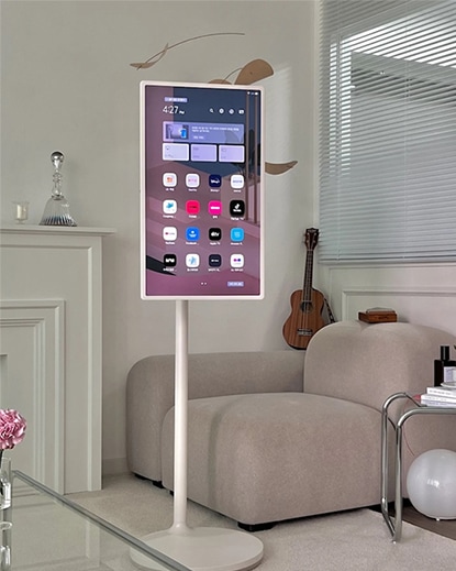 LG 스탠바이미가 거실 한가운데 자유롭게 배치되어 있으며, 스마트 인터페이스 화면을 통해 다양한 콘텐츠를 사용할 수 있는 모습을 보여줍니다. 화면 회전 및 각도 조절이 가능한 유연한 디자인으로 공간 활용도를 높입니다.