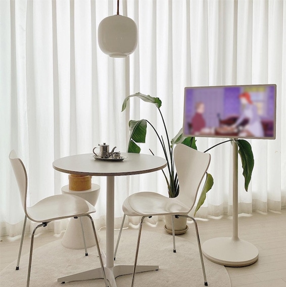 LG 스탠바이미가 밝은 커튼이 있는 창가 근처에 배치된 모습. TV 화면에는 흐릿한 영상이 표시되어 있으며, 옆에는 화분과 테이블, 그리고 화이트 컬러의 의자 두 개가 배치되어 깔끔하고 아늑한 분위기를 연출.